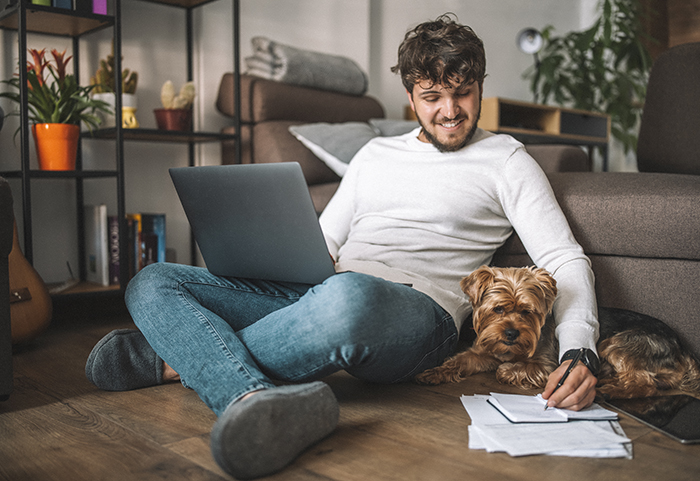 A shot of a person sitting on the floor with their laptop and smiling, while dog sleeps next to them