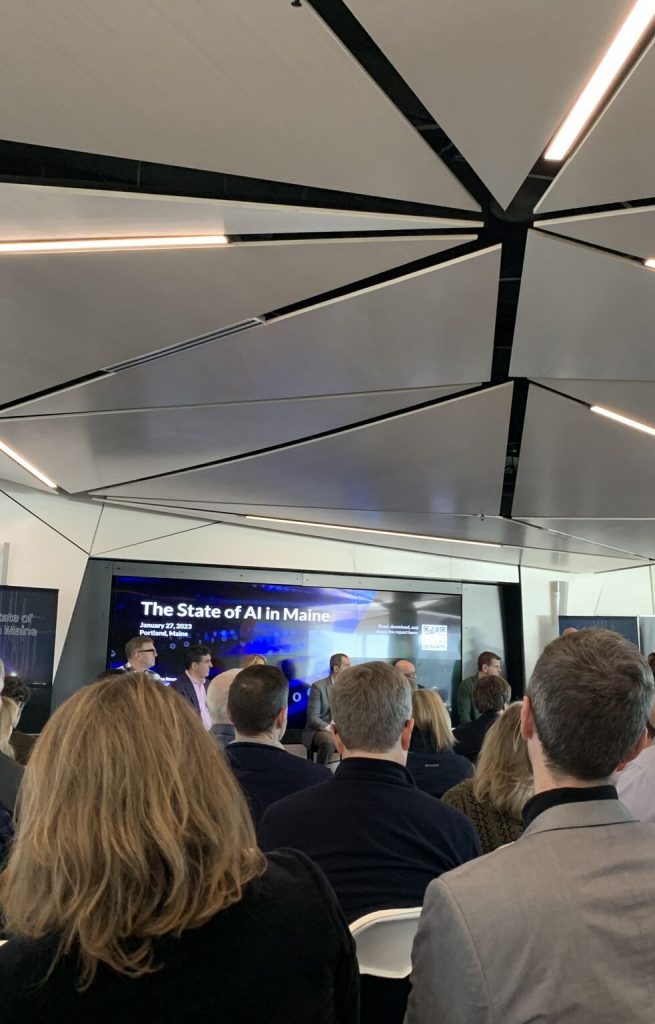 A crowd of attendees watches a panel of speakers being introduced at an A I event.