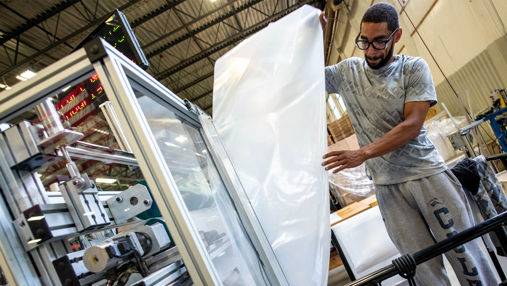 Worker operating machinery in a manufacturing facility, inspecting a large sheet of plastic film being processed through an industrial machine. The environment features high ceilings, metal framework, and visible equipment.