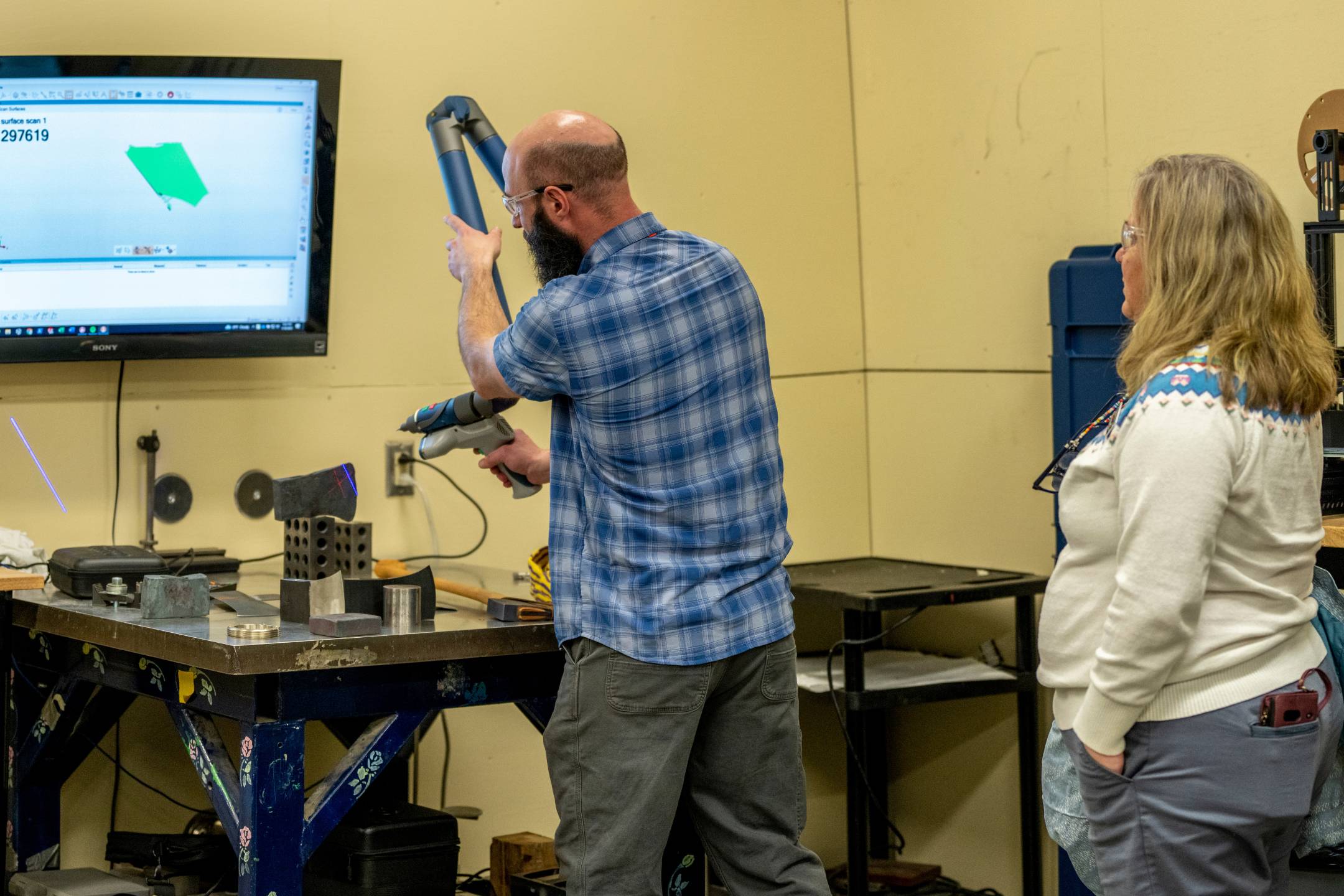 An Advanced Manufacturing Center staff member demonstrates a handheld 3D scanner while capturing a part’s surface geometry, with the live scan displayed on a monitor during a facility walkthrough.