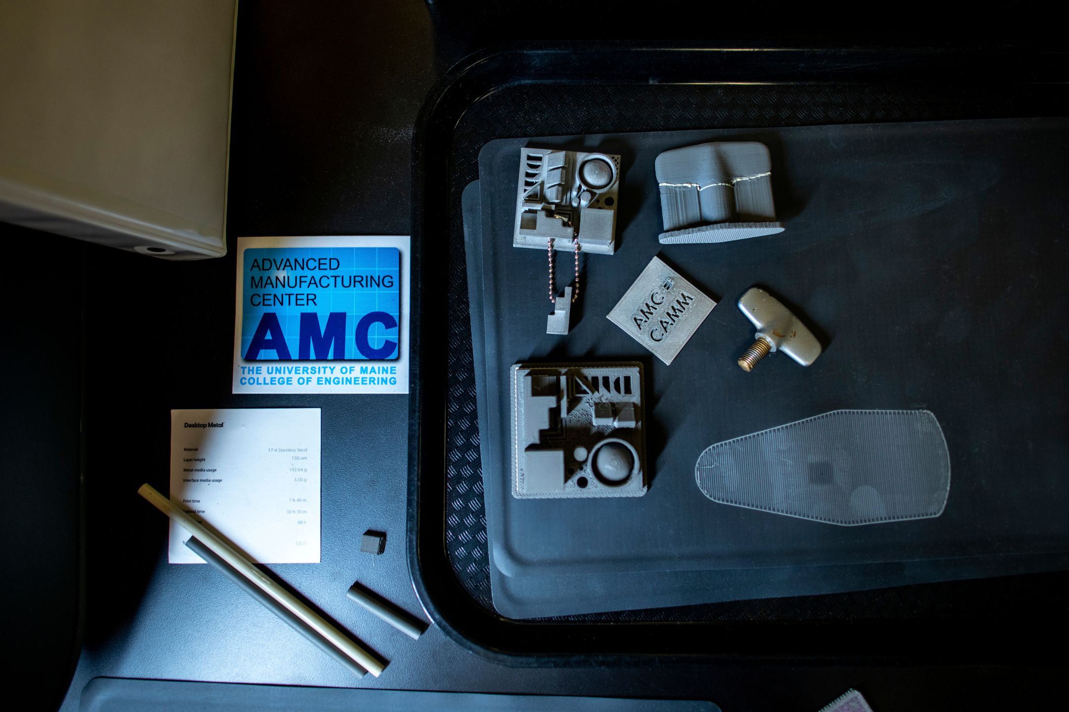 A collection of metal and polymer 3D-printed prototypes displayed next to an Advanced Manufacturing Center logo at the University of Maine, showcasing parts produced through additive manufacturing and CAM processes.