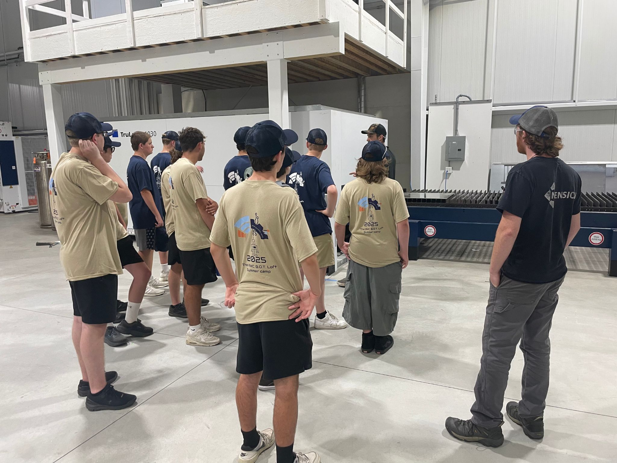 Campers in matching B.O.T. Loft shirts listen to a technician explain equipment during a guided tour of Ntension Corporation, observing a large industrial machine inside the facility.
