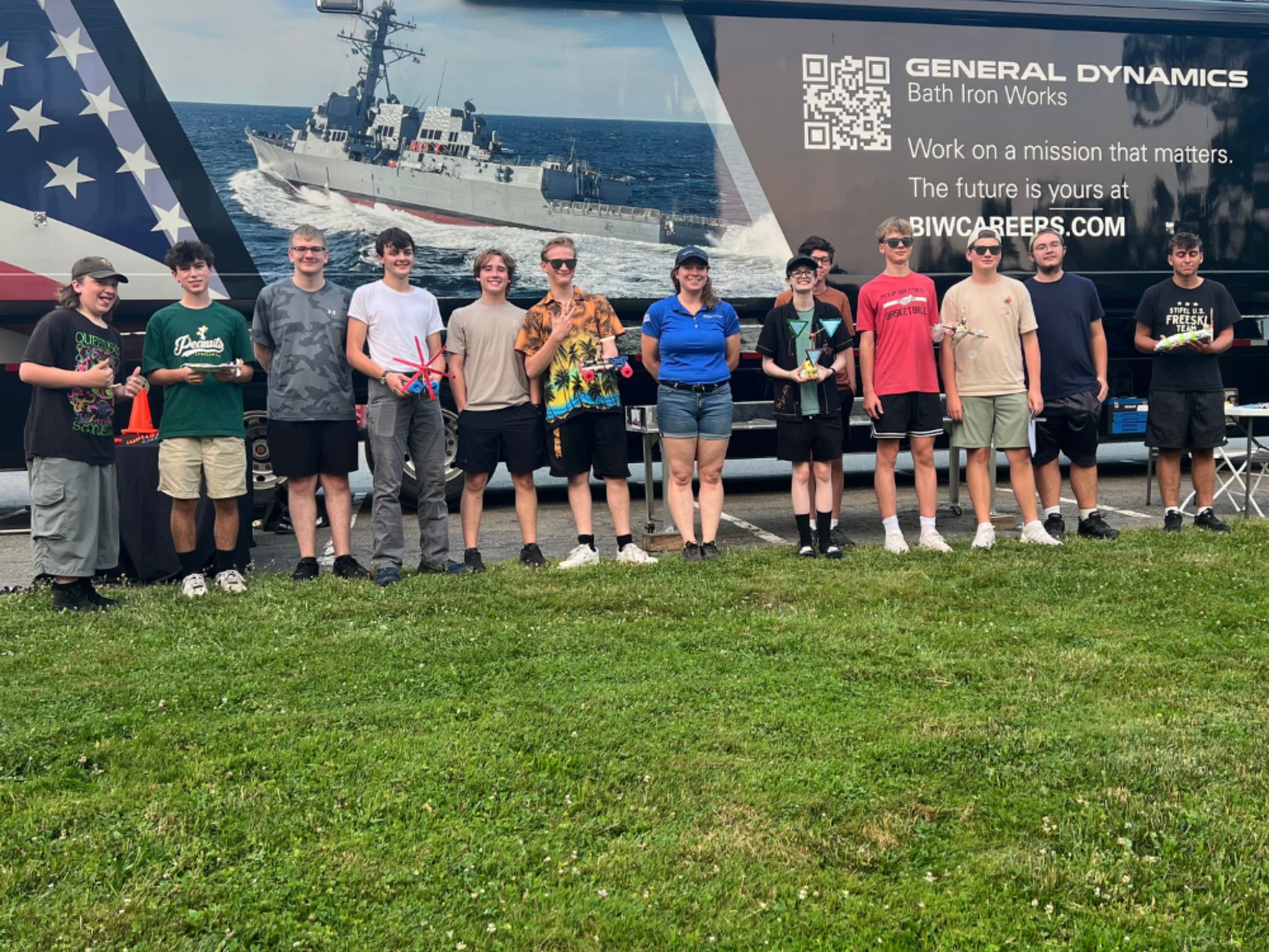 B.O.T. Loft campers pose proudly with their completed engineering projects in front of the Bath Iron Works mobile welding lab. The backdrop features a naval ship and recruitment messaging from General Dynamics, highlighting STEM career pathways.