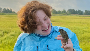 Woman with red hair in light blue jacket holds a cute little guy, a little saltmarsh sparrow.