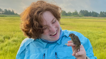 Woman with red hair in light blue jacket holds a cute little guy, a little saltmarsh sparrow.