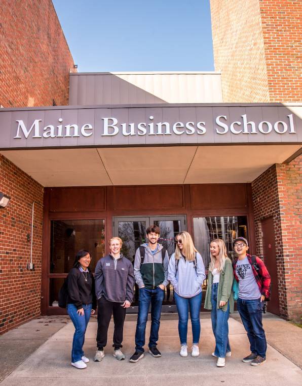 Image of six students standing in front of the DP Corbett building entrance on the Orono Maine campus of the Maine Business School