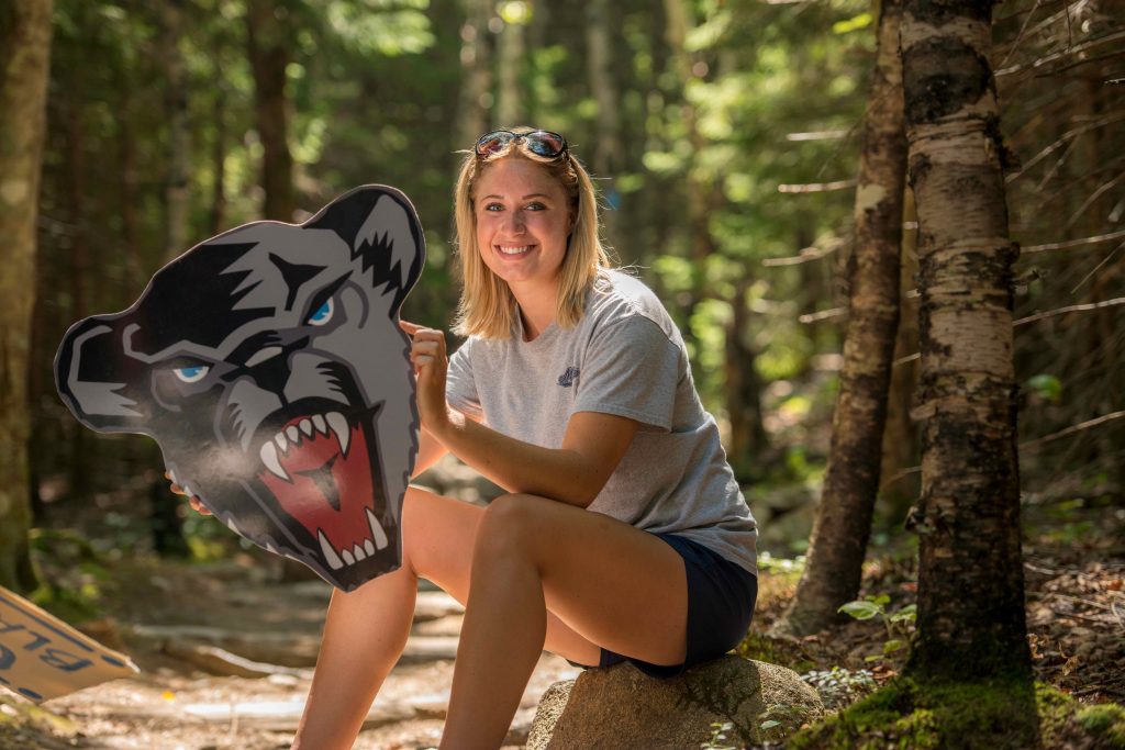 A photo of a student seated in the woods, holding a UMaine Black Bears mascot sign