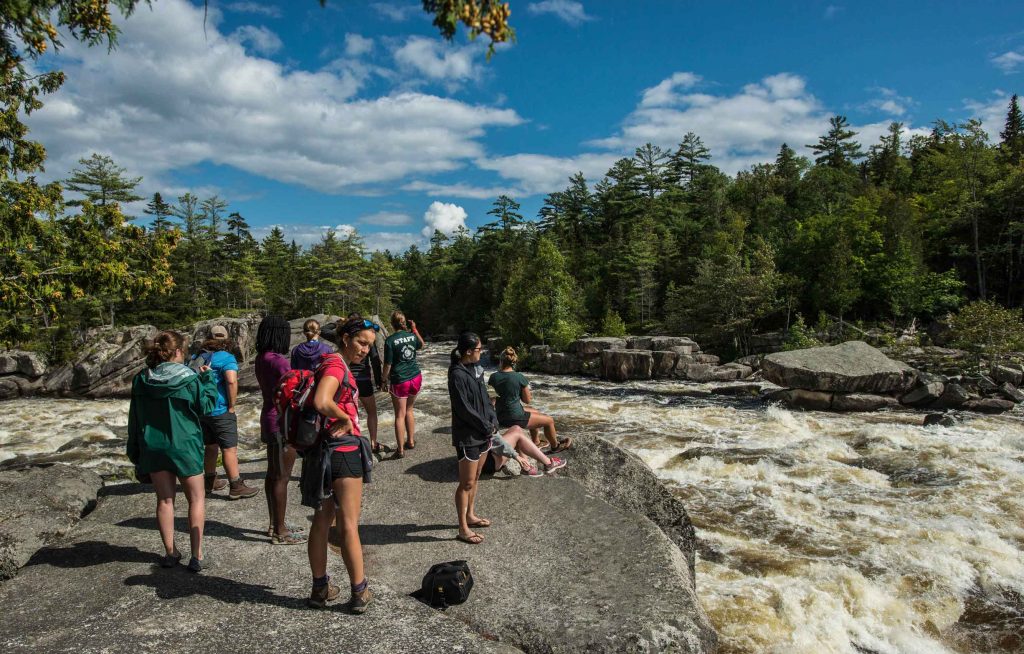 A group of students stand on a large flat rock with water rapids flowing around them