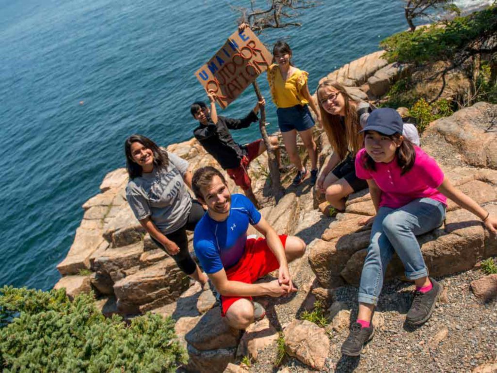 Students on the rocky coast of maine, one holding a sign reading Outdoor Nation