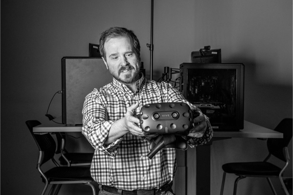 A black and white photo of Dr. Justin Dimmel holding a virtual reality headset with computer equipment in the background. 