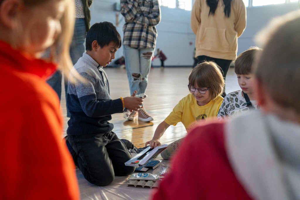 A small group of children working on an activity together in Lengyel Gym, while being observed by UMaine students in the background. 