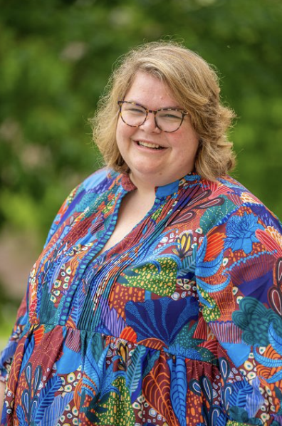 A photo of Dr. Catharine Biddle smiling. She is wearing glasses and a colorful, patterned blouse, standing outdoors with green foliage in the background