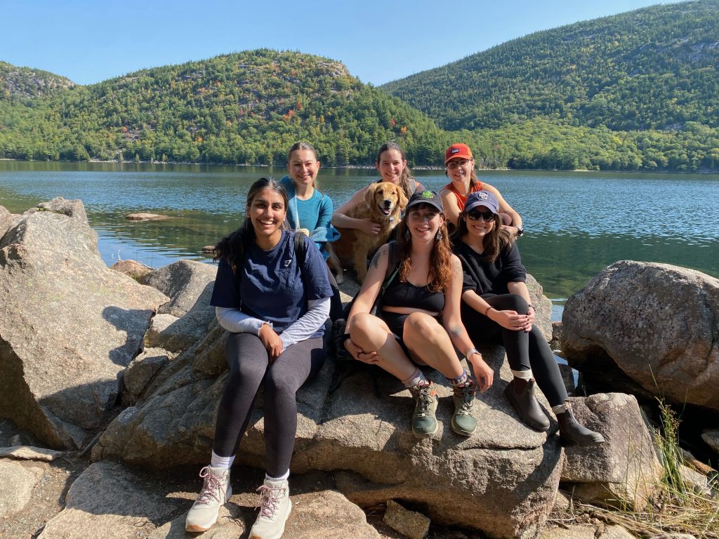 The 6 C-CAP Lab students (Caroline Kelberman, Eleanor Schuttenberg, Hannah Milius, Katie Guajardo, Aurora Green, and Puneet Parmar) and Lexie the Labrador dog sitting on some rocks in front of Jordan Pond at Acadia National Park.