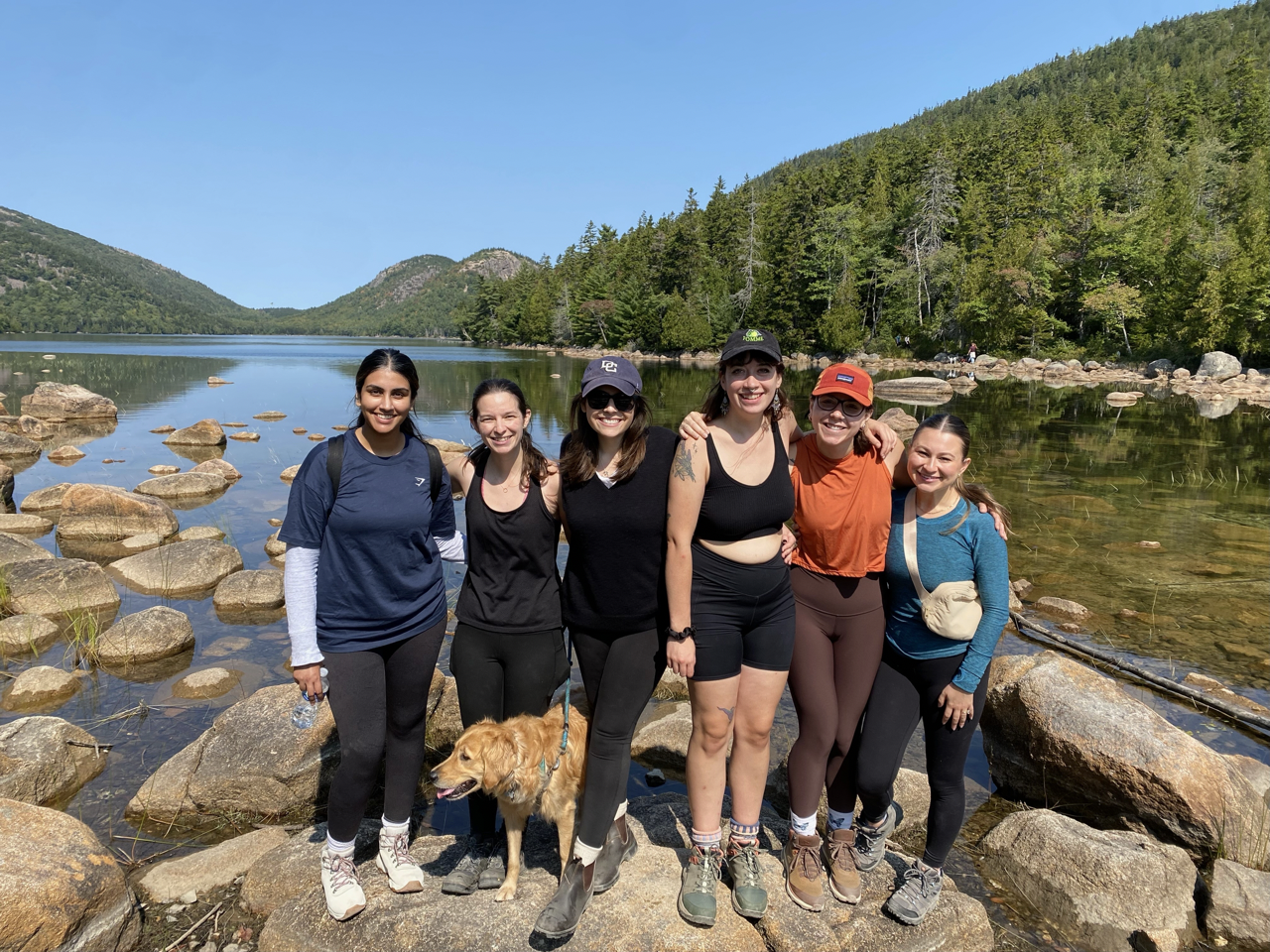 The 6 C-CAP Lab students (Caroline Kelberman, Eleanor Schuttenberg, Hannah Milius, Katie Guajardo, Aurora Green, and Puneet Parmar) and Lexie the Labrador dog sitting on some rocks in front of Jordan Pond at Acadia National Park.