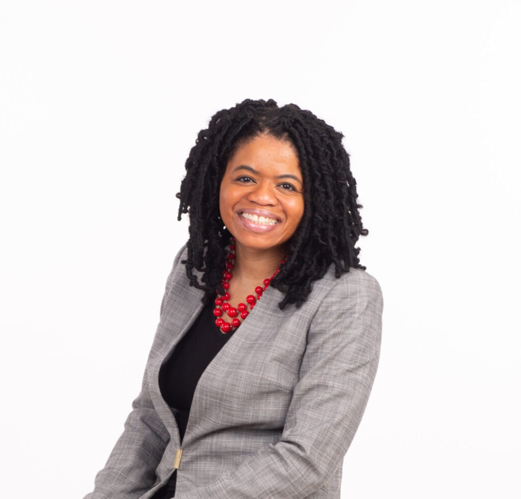 A photo of Dr Tracie Addie seated at a chair and smiling towards the camera. She is wearing a grey jacket, red necklace and black shirt.