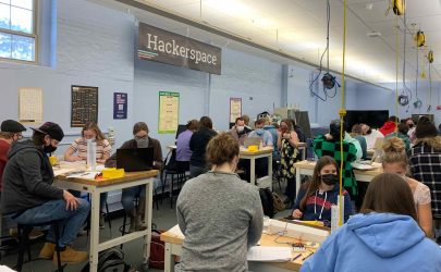 Numerous students are sitting around elevated work stations. Computers and other electronics are on the tables. A banner with the word Hackerspace hangs on the wall