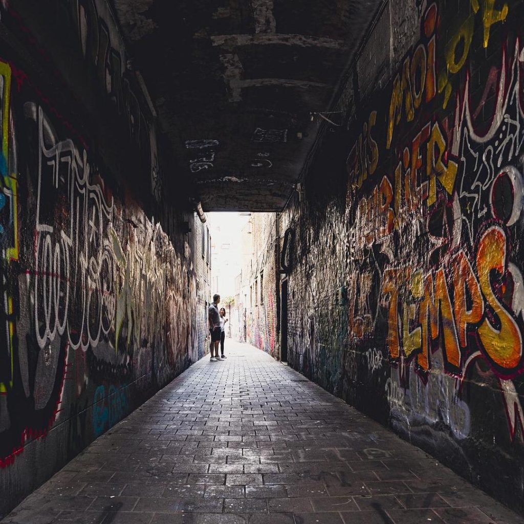 Covered path in Gent, Belgium. Walls and ceiling are covered in Graffiti. Two people are standing at the end of the path where it meets a narrow street in the daylight.