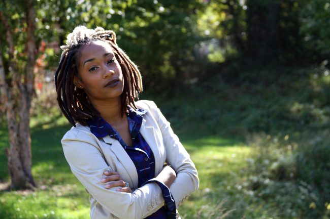 3/4 portrait of a black individual with shoulder length dreadlocks. dark brown eyes, and small earrings, wearing a cream colored suit jacket and a blue buttoned shirt featuring well spaced red vertical lines and white horizontal lines, looking at the camera with their arms crossed on their chest.