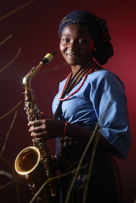 3/4 portrait of a young dark skinned woman wearing a black turban with gold motifs, a blue top with short sleeves and a red necklace and bracelet set, smiling at the camera while holding a saxophone, ready to perform.
