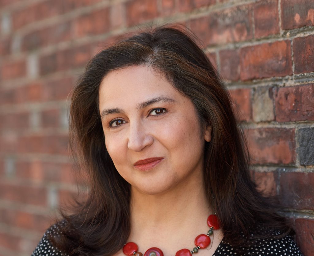 Photograph of Dr. Rajika Bhandari standing against a brick wall, smiling at the cameara with a red necklace.