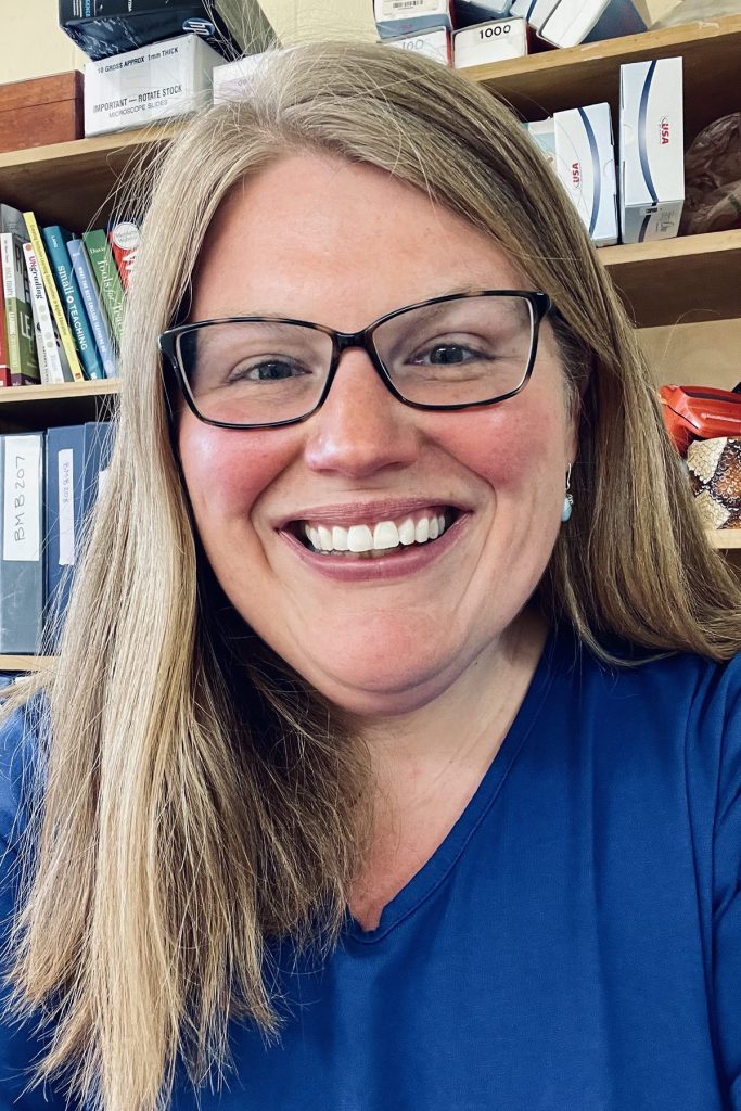 Headshot of professor Newell-Caito in a blue shirt and wearing square glasses positioned in front of a book shelf.