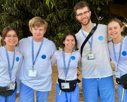 Five volunteer healthcare students stand together, smiling. They all wear white shirts with blue dots, blue scrubs, and ID badges. They look ready for work outdoors, standing under a tree.