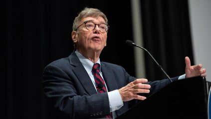 Charlie Cook, wearing glasses and a suit with a red and blue striped tie, speaks at a podium. He gestures with his hands as he addresses the audience, with a microphone in front of him and a black curtain as the backdrop.