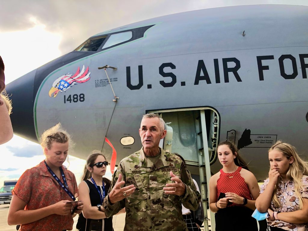 An officer in camouflage uniform stands before the head of a U.S. Air Force plane, leading a talk. Students gather around and listen.  