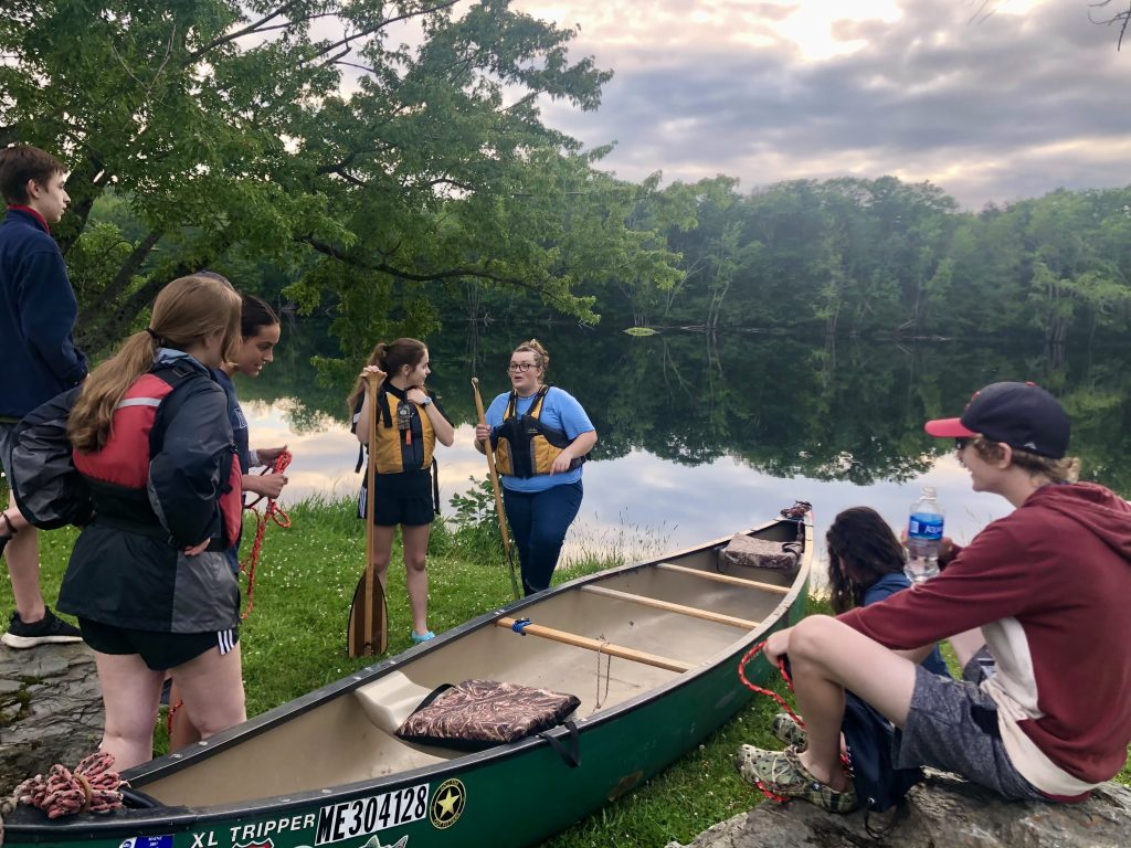 Students gather around a leader with a lifejacket and a paddle, who discusses protocol for launching the canoe pulled up on the grass in the center of the group. A calm river reflects the forest and sky in the background. 