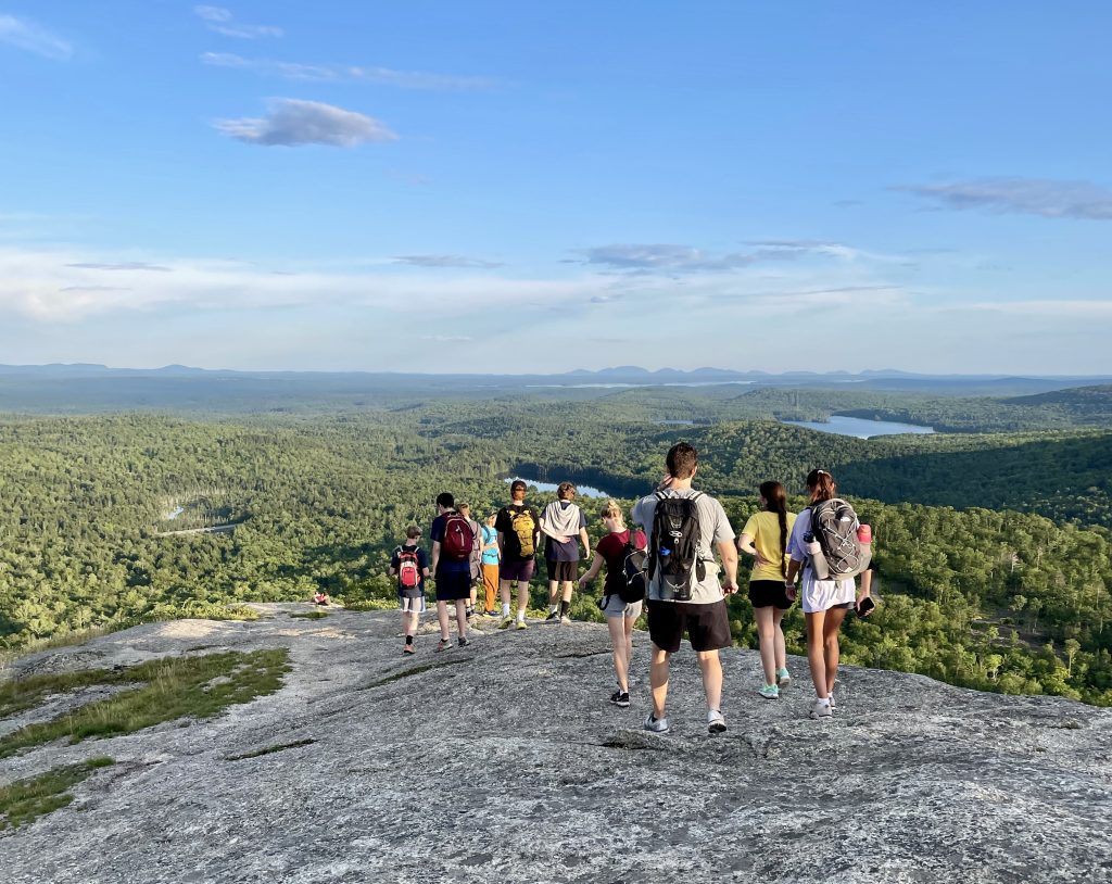 Leadership students hike in a group down a broad rocky mountain outcrop with the Maine wilderness in the vista below. 