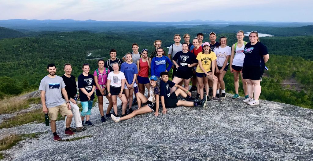 A group of leadership students stand and sit on a rocky outcrop, overlooking a scenic, forested landscape with distant mountains. The group appears to be relishing the natural surroundings and the panoramic view, showcasing a moment of camaraderie and adventure in a picturesque setting.