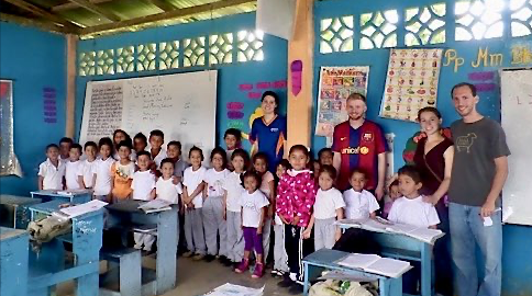 A brightly lit classroom with blue walls decorated with colorful educational posters and charts. The atmosphere is lively and vibrant. Children in white shirts and gray pants, along with casually dressed adults, are gathered around desks and benches filled with papers.