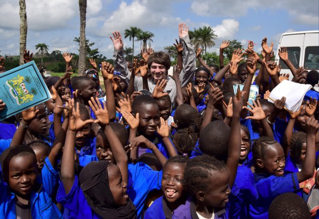 A large group of children wearing blue uniforms gather outdoors with their hands raised enthusiastically. Behind them are palm trees and a partly cloudy sky. In the center of the group, a University of Maine student joins in with a big smile.