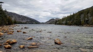 A view of one of the lakes in Acadia National Park with mountains in the background, pine trees lining the shore that recedes into the distance, and large rocks protruding from the shallow water in the foreground of the photos