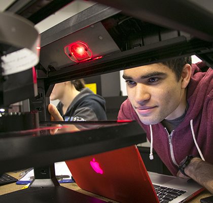 A student looks through the open center to the print surface of a 3D extrusion printer.