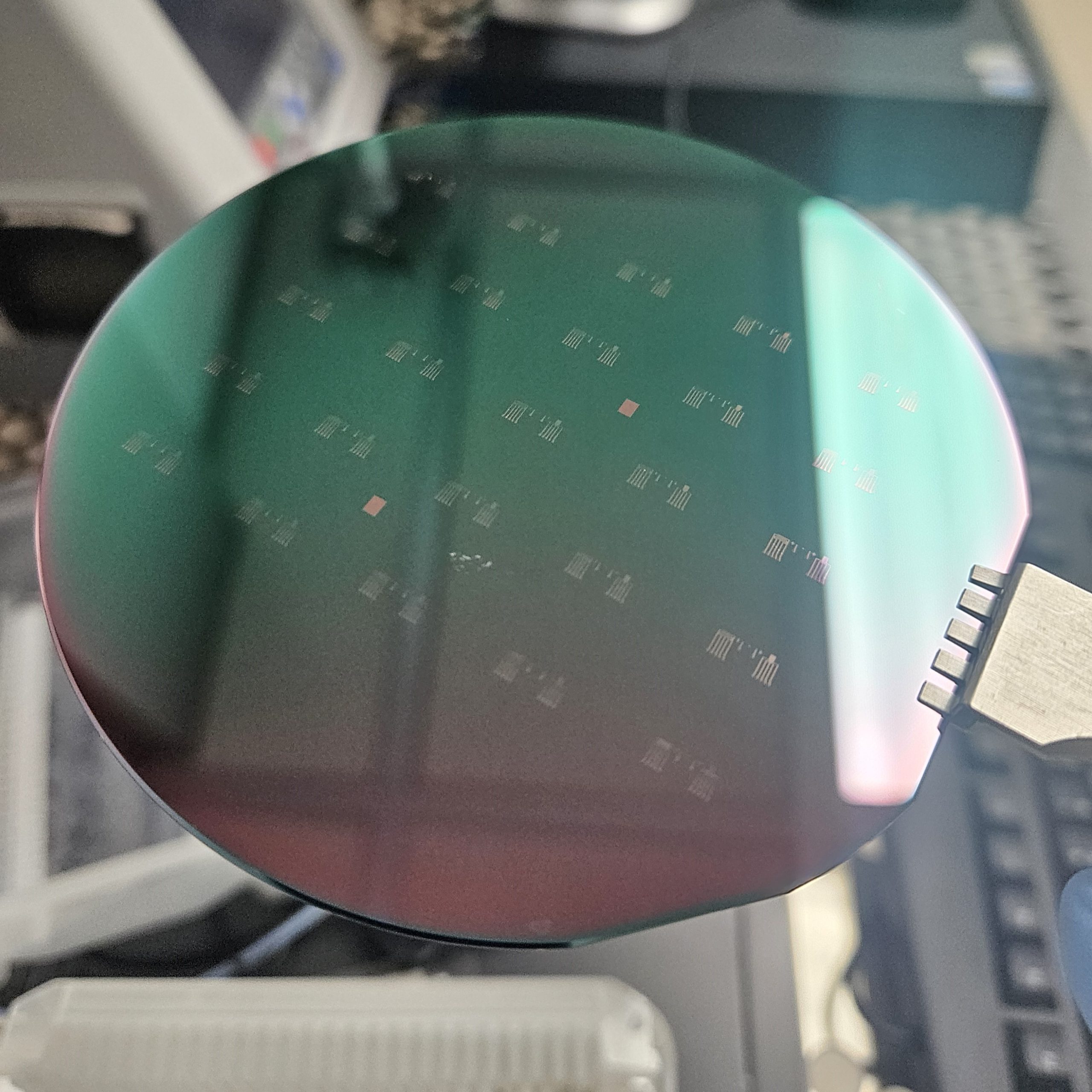 Michael Sabol's photograph of a microfabrication wafer. The resist layer baked on to the round wafer appears to have a green and pink reflection. Small electrodes can be seen printed on the wafer in a regular pattern.