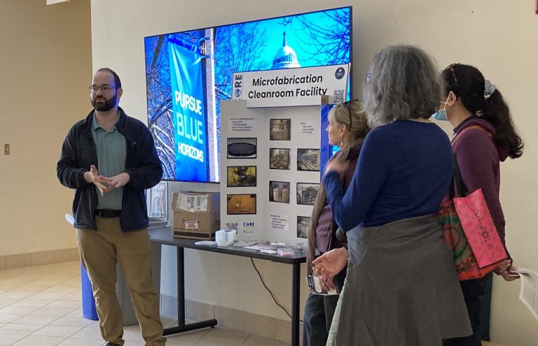 Carfagno stands at the left of the image, in front of a table with a trifold display. Several other people stand to the right of the table, listening.