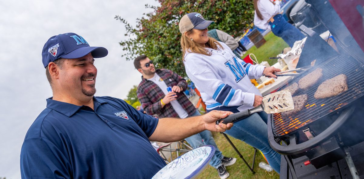 man grilling hamburgers and woman in the back with UMaine apparel on