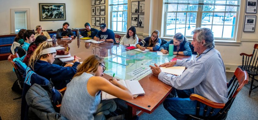 class sitting around long wooden table with professor seated at the head of the table