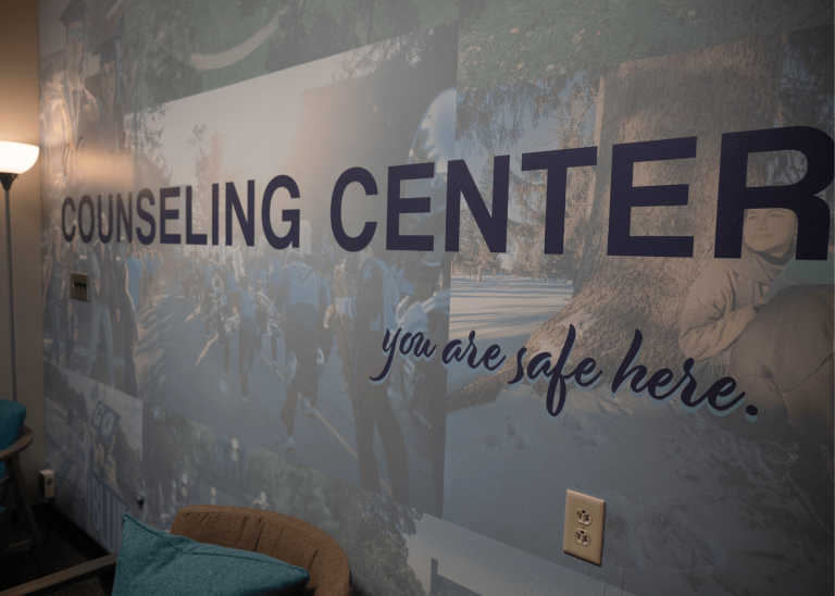 Wall of counseling center that says "counseling center, you are safe here" with collage of students as the background.