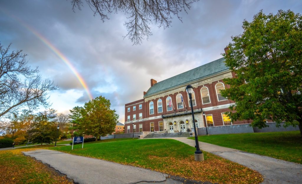 A photograph showing the front steps of Fogler Library, with a cloudy sky. A rainbow is visible on the left. Scattered orange leaves can be seen on the ground.