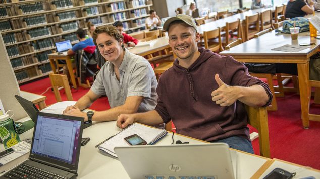 two guys with computers in library smiling, one giving a thumbs up