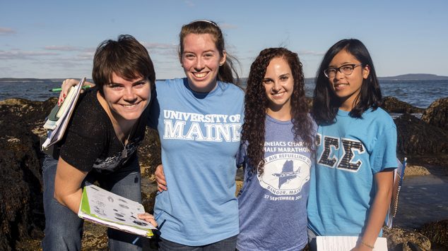 four students smiling and holding schoolwork by the ocean