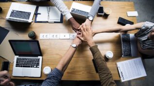 Group of hands in the middle of a table covered with laptops and paper