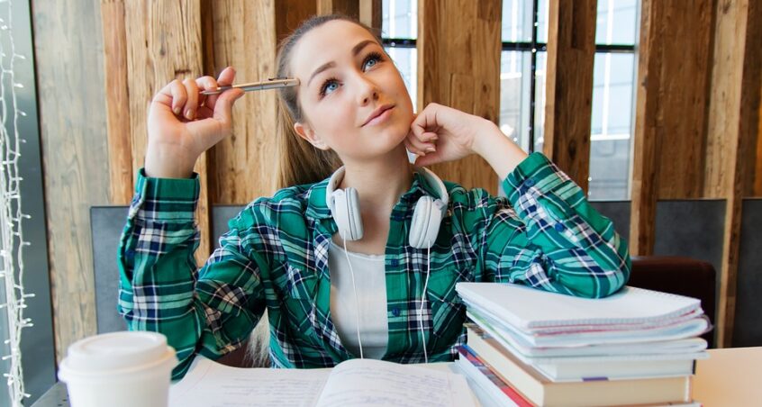 student looking off thoughtfully with a pen in her hand. She is sitting in front of a stack of books, a cup of coffee, and an open notebook.