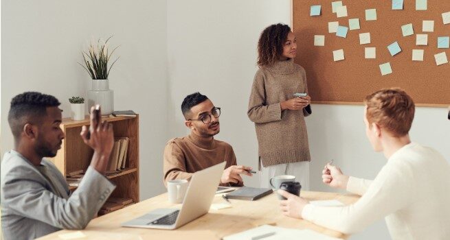 Tutor and students sit around a table with a corkboard on the wall and a laptop on the table