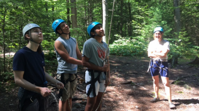 Students wearing climbing gear and helmets look up into the rope course.