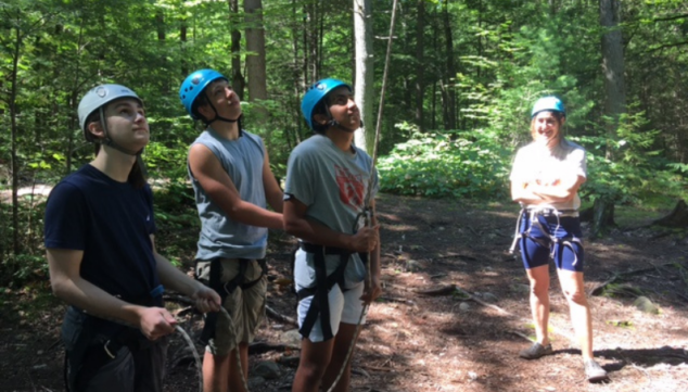 Students wearing climbing gear and helmets look up into the rope course.