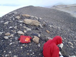 Calvin Mako takes notes near a basalt cobble collected for surface exposure dating. Calvin and the cobble are perched on the crest of a moraine ridge deposited by the Ross Sea ice sheet at the last glacial maximum. The view is to the Southwest, with Heald island and the Koettlitz glacier in the upper left background.