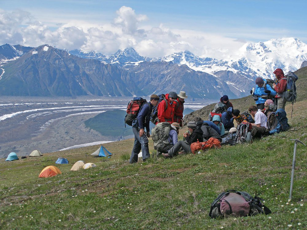 JIRP students looking at samples on a grassy hill, with a mountain landscape behind them.
